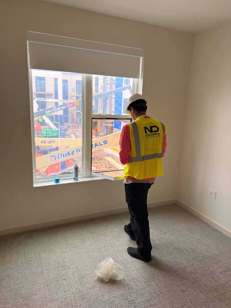 ND Cleaning crew member in a hard hat and safety vest performing final construction window cleaning in a newly finished room